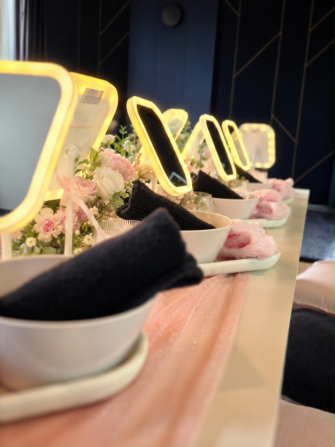 Side view of a styled Mini Luxe Spa Party table with illuminated mirrors, white bowls, navy towels and pink spa accessories.